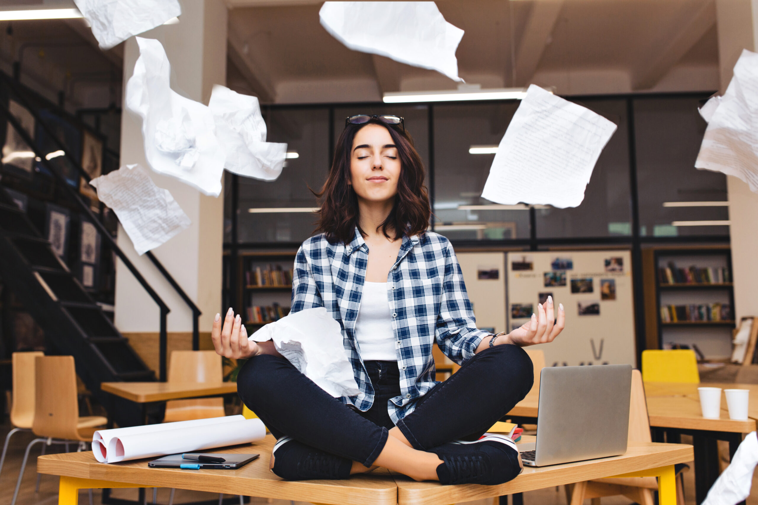 Young pretty joyful brunette woman meditating on table surround work stuff and flying papers. Cheerful mood, taking a break, working, studying, relaxation, true emotions